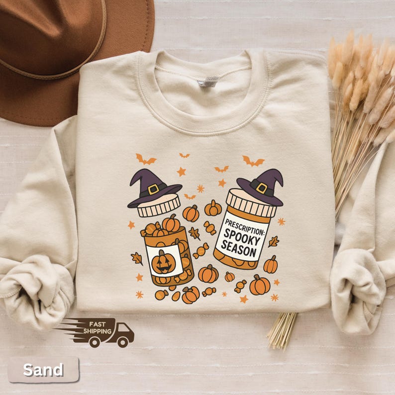 Op de afbeelding: Zandkleurige sweatshirt met een Halloween-thema. Het ontwerp toont twee medicijnflesjes met heksenhoeden, pompoenen en de tekst "Prescription: Spooky Season". Vleermuizen en sterren maken de feestelijke look compleet.