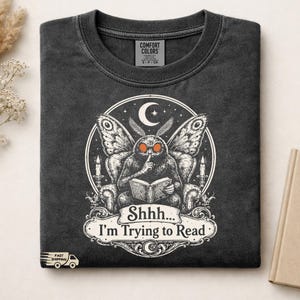 May include: A charcoal gray t-shirt featuring a graphic of a moth sitting in an armchair reading a book with a finger to its mouth. The design includes a crescent moon, stars, and two candles. The text reads, Shhhh... I'm Trying to Read.