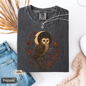 May include: Dark grey Comfort Colors t-shirt with an owl illustration, crescent moon, and floral wreath design. A silver necklace with a sun pendant rests on the shirt. The shirt is folded on a white surface with blue jeans.