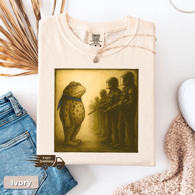 K&ouml;nnte beinhalten: Elfenbeinfarbenes T-Shirt mit einem Cartoon-Frosch mit blauem Schal, der Figuren mit Helmen und Waffen gegen&uuml;bersteht. Die Grafik ist gerahmt. Das Shirt hat ein Comfort Colors-Etikett. Das Wort "Ivory" ist aufgedruckt.
