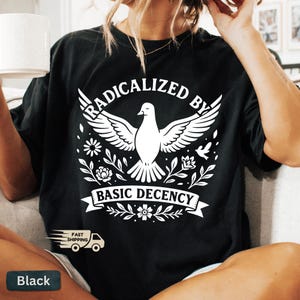 Op de afbeelding: Zwart T-shirt met een witte afbeelding van een duif met gespreide vleugels, omringd door bloemen. De tekst "RADICALIZED BY BASIC DECENCY" staat erboven en eronder. Inclusief een "Snelle verzending"-logo.