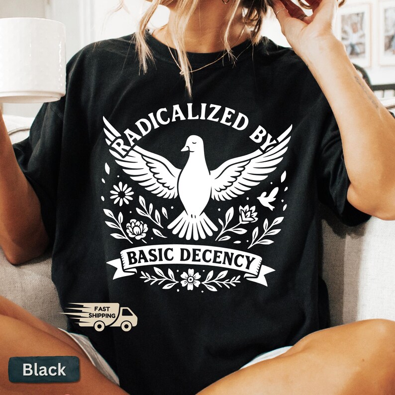 Op de afbeelding: Zwart T-shirt met een witte afbeelding van een duif met gespreide vleugels, omringd door bloemen. De tekst "RADICALIZED BY BASIC DECENCY" staat erboven en eronder. Inclusief een "Snelle verzending"-logo.