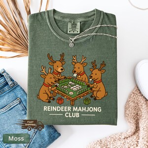 Puede incluir: Camiseta verde musgo con una ilustraci&oacute;n de dibujos animados de cuatro renos jugando al mahjong. El texto "REINDEER MAHJONG CLUB" est&aacute; impreso debajo. Un collar plateado est&aacute; sobre la camiseta.