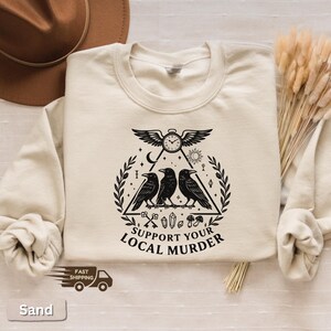 May include: Sand-colored sweatshirt with a black graphic featuring three crows inside a triangle, a clock, and the text "SUPPORT YOUR LOCAL MURDER." The design includes keys, crystals, and mushrooms. The sweatshirt has a crew neck and long sleeves.