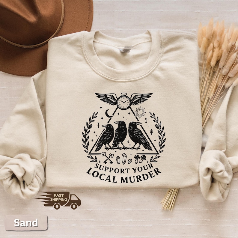 May include: Sand-colored sweatshirt with a black graphic featuring three crows inside a triangle, a clock, and the text "SUPPORT YOUR LOCAL MURDER." The design includes keys, crystals, and mushrooms. The sweatshirt has a crew neck and long sleeves.