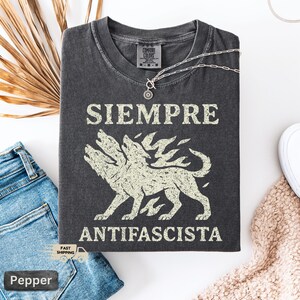 May include: Dark grey t-shirt with the words "SIEMPRE ANTIFASCISTA" in white block letters above and below a three-headed wolf illustration. The shirt is folded on a white surface next to blue jeans and a necklace.
