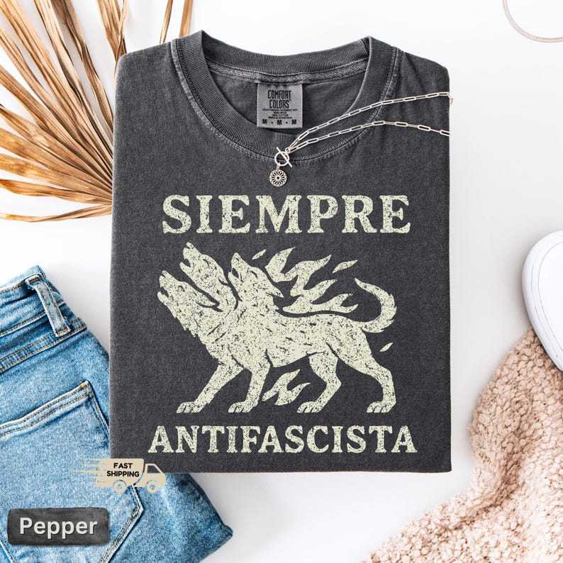 May include: Dark grey t-shirt with the words "SIEMPRE ANTIFASCISTA" in white block letters above and below a three-headed wolf illustration. The shirt is folded on a white surface next to blue jeans and a necklace.