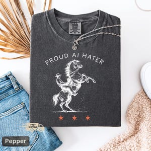 May include: Dark gray Comfort Colors t-shirt with the text "PROUD AI HATER" and a white illustration of a cowboy riding a rearing horse. The shirt also features three orange stars at the bottom. A silver necklace with a sun pendant is draped across the shirt.