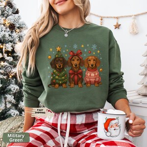 May include: Military green sweatshirt featuring three dachshunds dressed for Christmas. One dog is in a Christmas tree costume, another with a red bow, and the third in a sweater. The words "Fast Shipping" are printed on the left side of the sweatshirt.