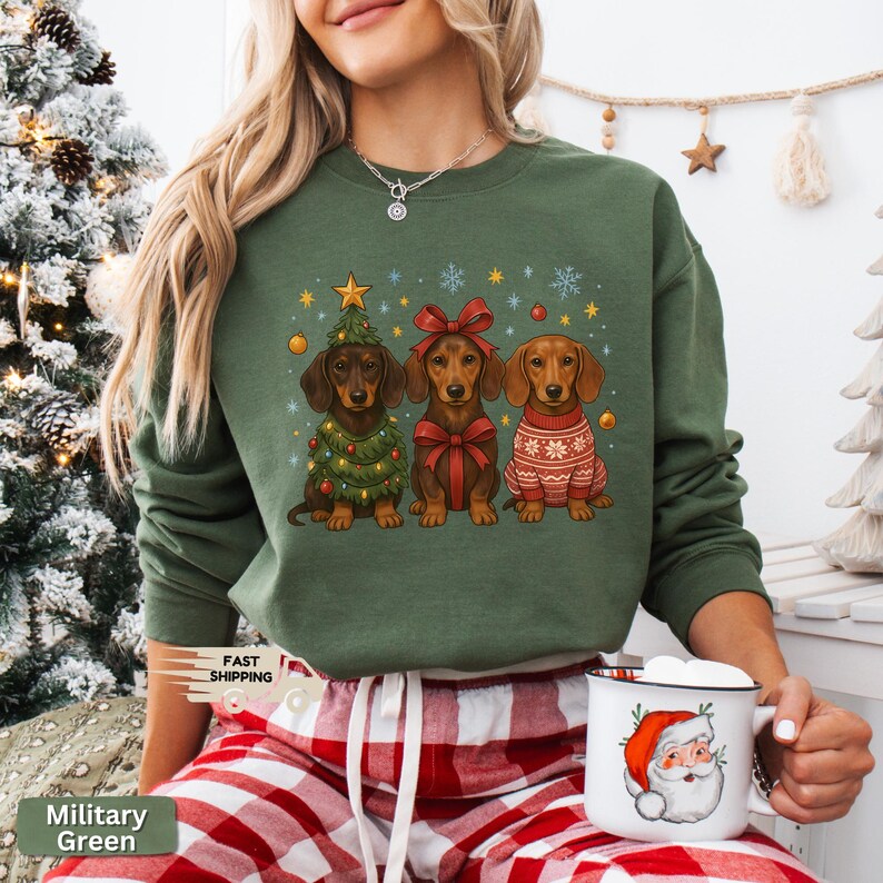 May include: Military green sweatshirt featuring three dachshunds dressed for Christmas. One dog is in a Christmas tree costume, another with a red bow, and the third in a sweater. The words "Fast Shipping" are printed on the left side of the sweatshirt.