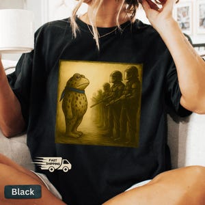 K&ouml;nnte beinhalten: Schwarzes T-Shirt mit einem quadratischen Grafikdesign, das einen Frosch mit blauem Schal zeigt, der Soldaten gegen&uuml;bersteht. Der Frosch ist hellbraun mit dunklen Flecken. Die Soldaten tragen dunkle Uniformen.