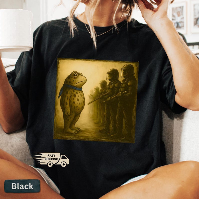 K&ouml;nnte beinhalten: Schwarzes T-Shirt mit einem quadratischen Grafikdesign, das einen Frosch mit blauem Schal zeigt, der Soldaten gegen&uuml;bersteht. Der Frosch ist hellbraun mit dunklen Flecken. Die Soldaten tragen dunkle Uniformen.