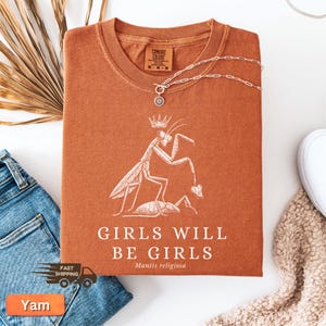 May include: A rust-colored t-shirt with a white graphic of a praying mantis wearing a crown, with the text "GIRLS WILL BE GIRLS" and "Mantis religiosa". A silver necklace with a pendant is draped across the shirt.