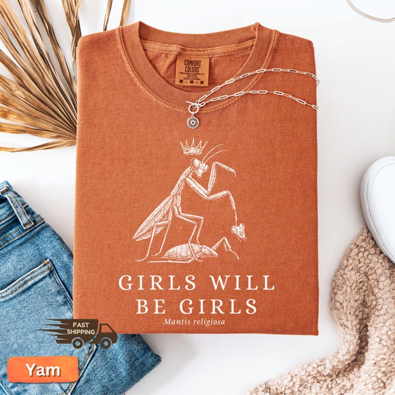 May include: A rust-colored t-shirt with a white graphic of a praying mantis wearing a crown, with the text "GIRLS WILL BE GIRLS" and "Mantis religiosa". A silver necklace with a pendant is draped across the shirt.