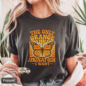 Op de afbeelding: Donkergrijze t-shirt met een monarchvlinderontwerp en de tekst "THE ONLY ORANGE MONARCH - I WANT-". De vlinder is oranje en zwart, tegen een gele en oranje achtergrond.