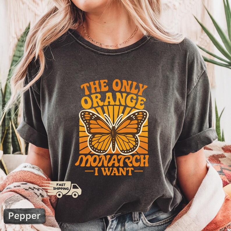 Op de afbeelding: Donkergrijze t-shirt met een monarchvlinderontwerp en de tekst "THE ONLY ORANGE MONARCH - I WANT-". De vlinder is oranje en zwart, tegen een gele en oranje achtergrond.