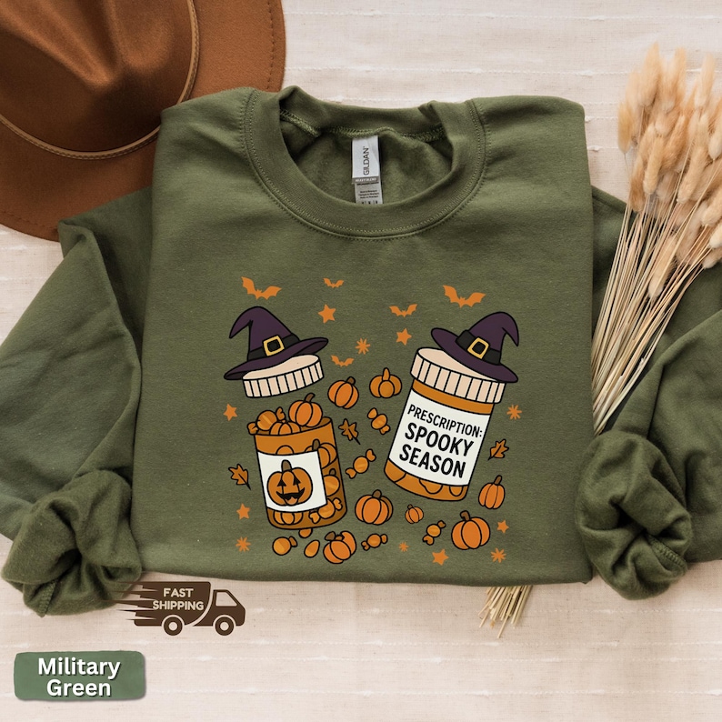 Op de afbeelding: Militairgroen sweatshirt met een Halloween-thema. Het ontwerp toont twee medicijnflesjes met heksenhoeden, pompoenen en de tekst "Prescription: Spooky Season". De trui heeft ook vleermuizen en sterren.