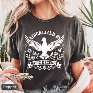 Op de afbeelding: Donkergrijs t-shirt met de witte tekst "RADICALIZED BY BASIC DECENCY". Het ontwerp toont een duif met uitgespreide vleugels, omringd door bloemen en bladeren. Het t-shirt heeft korte mouwen.
