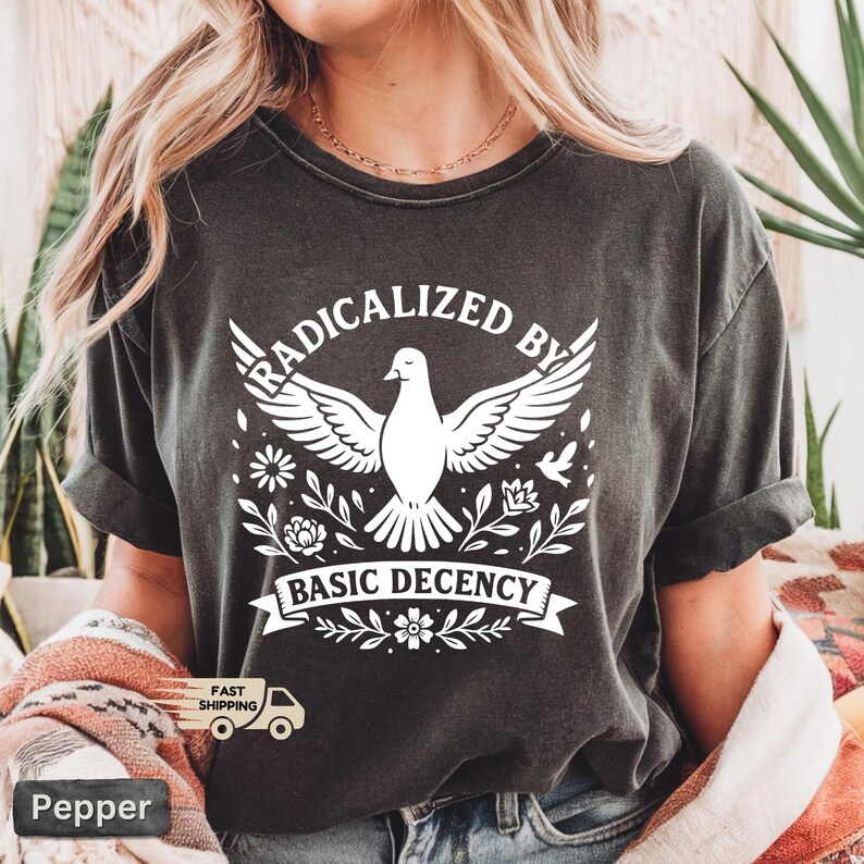Op de afbeelding: Donkergrijs t-shirt met de witte tekst "RADICALIZED BY BASIC DECENCY". Het ontwerp toont een duif met uitgespreide vleugels, omringd door bloemen en bladeren. Het t-shirt heeft korte mouwen.