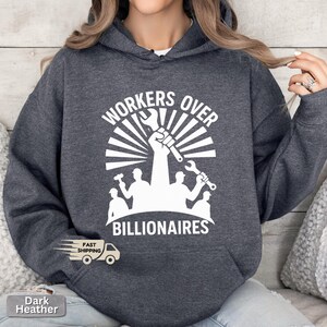 May include: Dark heather gray hoodie with the text "WORKERS OVER BILLIONAIRES" in white. The design features a raised fist holding a wrench, surrounded by a sunburst and silhouettes of workers with tools. The hoodie has a drawstring hood.