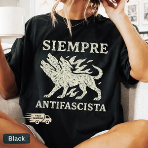May include: Black t-shirt with the words "SIEMPRE ANTIFASCISTA" in white, along with a three-headed dog graphic. The shirt has a vintage, distressed look. A small "Fast Shipping" logo is visible.