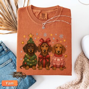May include: Rust-colored Comfort Colors t-shirt featuring three dachshunds. One dog is dressed as a Christmas tree, another with a red bow, and the third in a sweater. A silver necklace is draped on the shirt.
