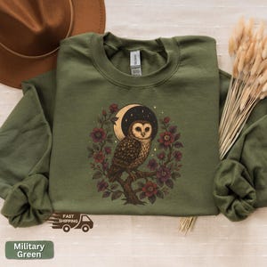 May include: Military green sweatshirt with an owl design, set against a crescent moon and starry sky, surrounded by burgundy flowers. The sweatshirt has long sleeves and a crew neck. The text "Military Green" is at the bottom.