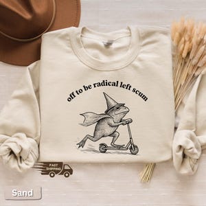 Pode incluir: Camisola cor de areia com a frase "off to be radical left scum" acima de um sapo de desenho animado andando de scooter. O sapo usa um chap&eacute;u de bruxa e capa. Gola redonda.
