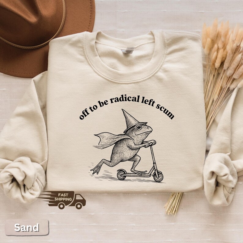 Pode incluir: Camisola cor de areia com a frase "off to be radical left scum" acima de um sapo de desenho animado andando de scooter. O sapo usa um chap&eacute;u de bruxa e capa. Gola redonda.