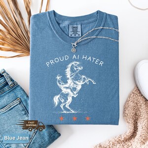 May include: A blue Comfort Colors t-shirt with the text "PROUD AI HATER" and a white graphic of a cowboy riding a rearing horse. A silver necklace with a pendant rests on the shirt. The shirt is folded, with blue jeans and a beige sweater visible.