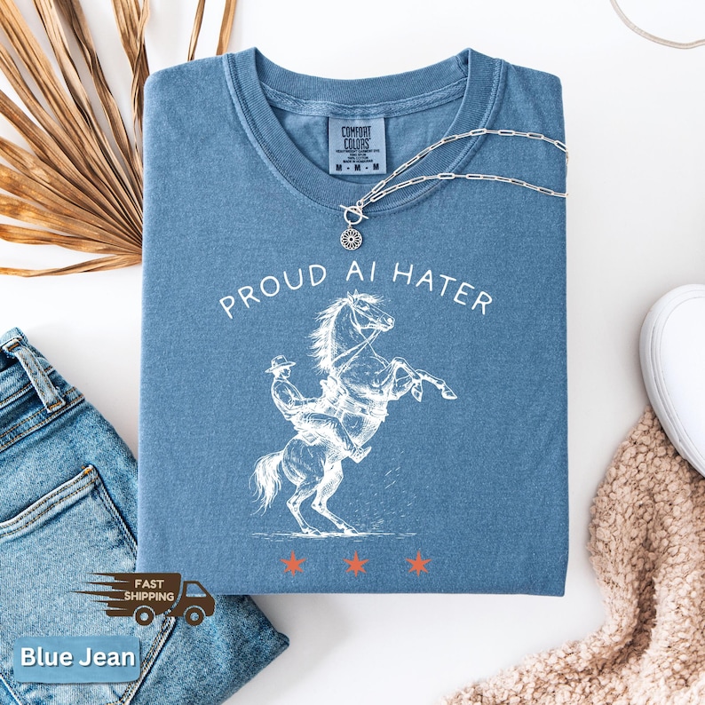 May include: A blue Comfort Colors t-shirt with the text "PROUD AI HATER" and a white graphic of a cowboy riding a rearing horse. A silver necklace with a pendant rests on the shirt. The shirt is folded, with blue jeans and a beige sweater visible.