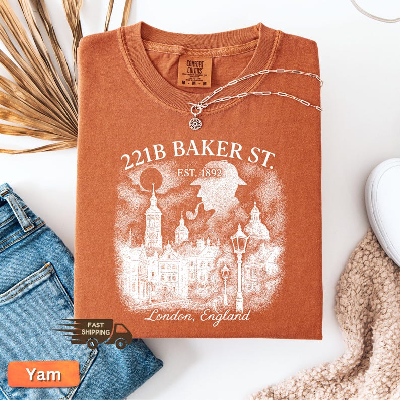 May include: A terracotta-coloured t-shirt with a white graphic that reads "221B BAKER ST. EST. 1892 London, England" and a silhouette of a person with a pipe and buildings. A silver necklace is draped across the shirt.
