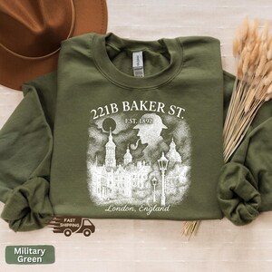 May include: A military green sweatshirt with a white graphic. The graphic includes the text "221B BAKER ST. EST. 1892 London, England" and a silhouette of a person with a pipe and a city scene.