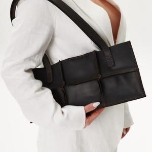 Può includere: Una borsa in pelle marrone scuro con forma rettangolare e design patchwork. La borsa ha una lunga tracolla marrone scuro. Una persona con un blazer bianco tiene la borsa.