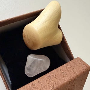 Peut inclure: Un objet en bois clair et lisse avec une forme ergonomique incurvée est placé dans une boîte cadeau brune texturée. À côté, une pierre polie rose pâle translucide repose sur un coussin en velours noir à l'intérieur de la boîte.