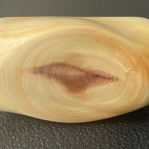 May include: A light-colored, wooden object with a curved shape. The wood grain is visible, with a darker, oval-shaped pattern in the center. The object appears to be smooth and polished, with rounded edges. The item is likely a decorative or functional wooden piece.