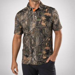 Può includere: Una polo a maniche corte con un realistico motivo mimetico boschivo nei toni del marrone, beige e verde. La maglia presenta un colletto con un'abbottonatura a tre bottoni, che rivela una fodera interna arancione brillante sul colletto.