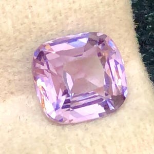 May include: A cushion-cut, light purple gemstone with facets. The gem is set against a white and dark green background, highlighting its color and clarity. The gemstone's shape is square with rounded corners.