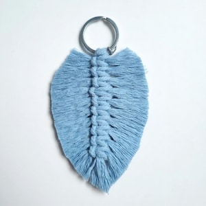 Può includere: Un portachiavi in macramè azzurro a forma di foglia, caratterizzato da una spina centrale annodata con bordi sfrangiati e un anello diviso in metallo argentato. Il testo Handmade 100% Cotton è visibile nell'angolo in basso a destra.