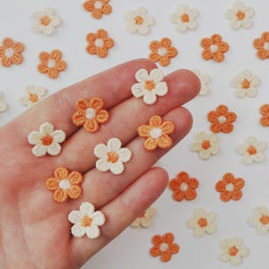 May include: Close-up of small, embroidered daisy flower patches in shades of orange and cream. The flowers are arranged on a white surface, with some held in a hand. These patches are ideal for crafting and embellishments.
