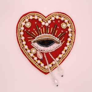 May include: A heart-shaped patch featuring red felt fabric decorated with gold, white, and black beads. The center displays an embroidered eye motif with radiating gold rays, surrounded by a border of white pearls and gold beads with dangling clear crystal-like teardrop accents.