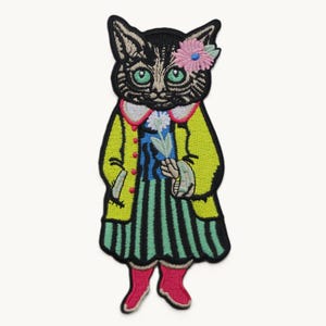 以下が含まれることがあります： 気まぐれな猫のキャラクターが特徴の刺繍パッチ。猫は黒とグレーの顔に緑の目、ピンクと青の花があり、ストライプのドレスの上に黄色のコートを着ています。猫は白い花を持っていて、ピンクのブーツを履いています。