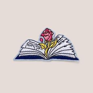 Puede incluir: Un parche bordado para planchar que presenta una rosa rosa creciendo de un libro abierto con páginas blancas y una cubierta azul.