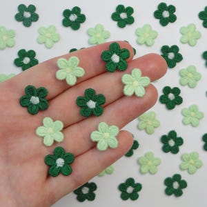 May include: Small, embroidered flower appliques in shades of green and light green. The appliques are in the shape of five-petal flowers with white centers. These decorative floral embellishments are ideal for craft projects.