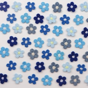Peut inclure: Une collection de petites appliques de fleurs crochetées dans diverses nuances de bleu, dont le marine, le ciel et le denim. Chaque fleur a un centre blanc et une touche de jaune. Les appliques sont disposées en grille.