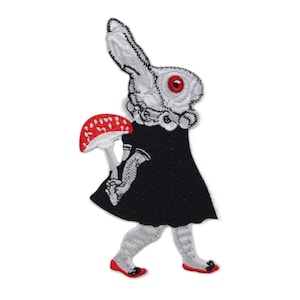 May include: Embroidered patch featuring a white rabbit with red eyes, wearing a black dress and holding a red and white mushroom. The rabbit has white and gray details, and is walking with red shoes. The patch is likely for clothing or accessories.