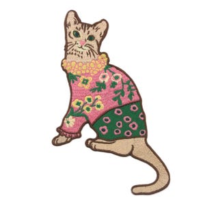 May include: Embroidered patch of a cat in a seated pose. The cat is wearing a pink sweater with floral embroidery and a green panel with white circles. The cat's collar is yellow, and the cat itself is tan.