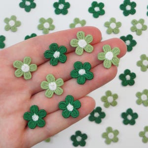 May include: A collection of small, embroidered flower appliques in shades of green and white. The flowers are in various shades of green, with white centers. The appliques are scattered across a surface, with some held in a hand.