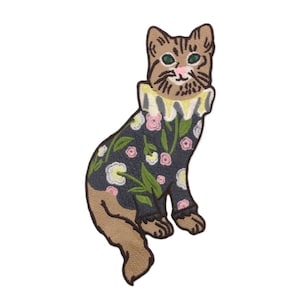 May include: Embroidered patch of a seated cat. The cat has a brown and tan body, green eyes, and a pink nose. It wears a dark blue outfit with a floral pattern of pink, yellow, and white flowers. The cat has a ruffled collar.