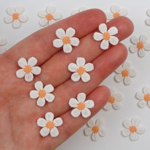 Op de afbeelding: Kleine witte madeliefjes bloem applicaties met oranje centra. Deze geborduurde stoffen bloemen zijn ideaal voor het verfraaien van kleding, accessoires of knutselprojecten. De madeliefjes hebben een diameter van ongeveer 2 cm.